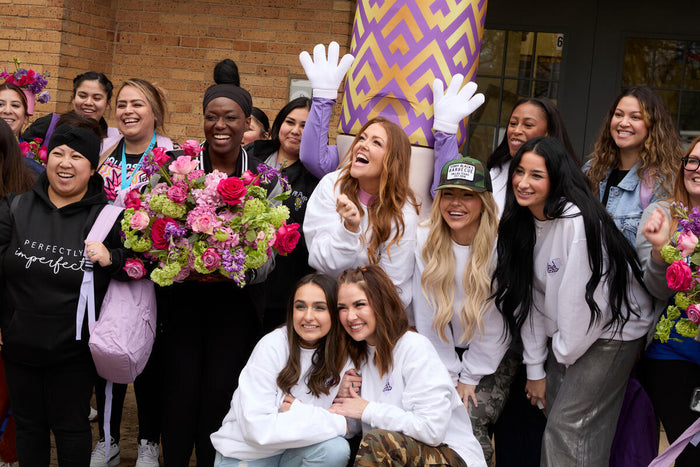 A group of people gathered, smiling and posing outdoors. One holds a bouquet of pink and purple flowers. Others are wearing casual clothing, standing near a brick building entrance.
