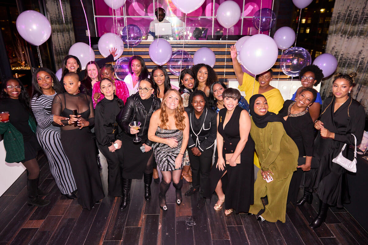 A group of people posing cheerfully with balloons in a festive indoor setting; behind them, a DJ stands at a booth with shiny purple decorations.