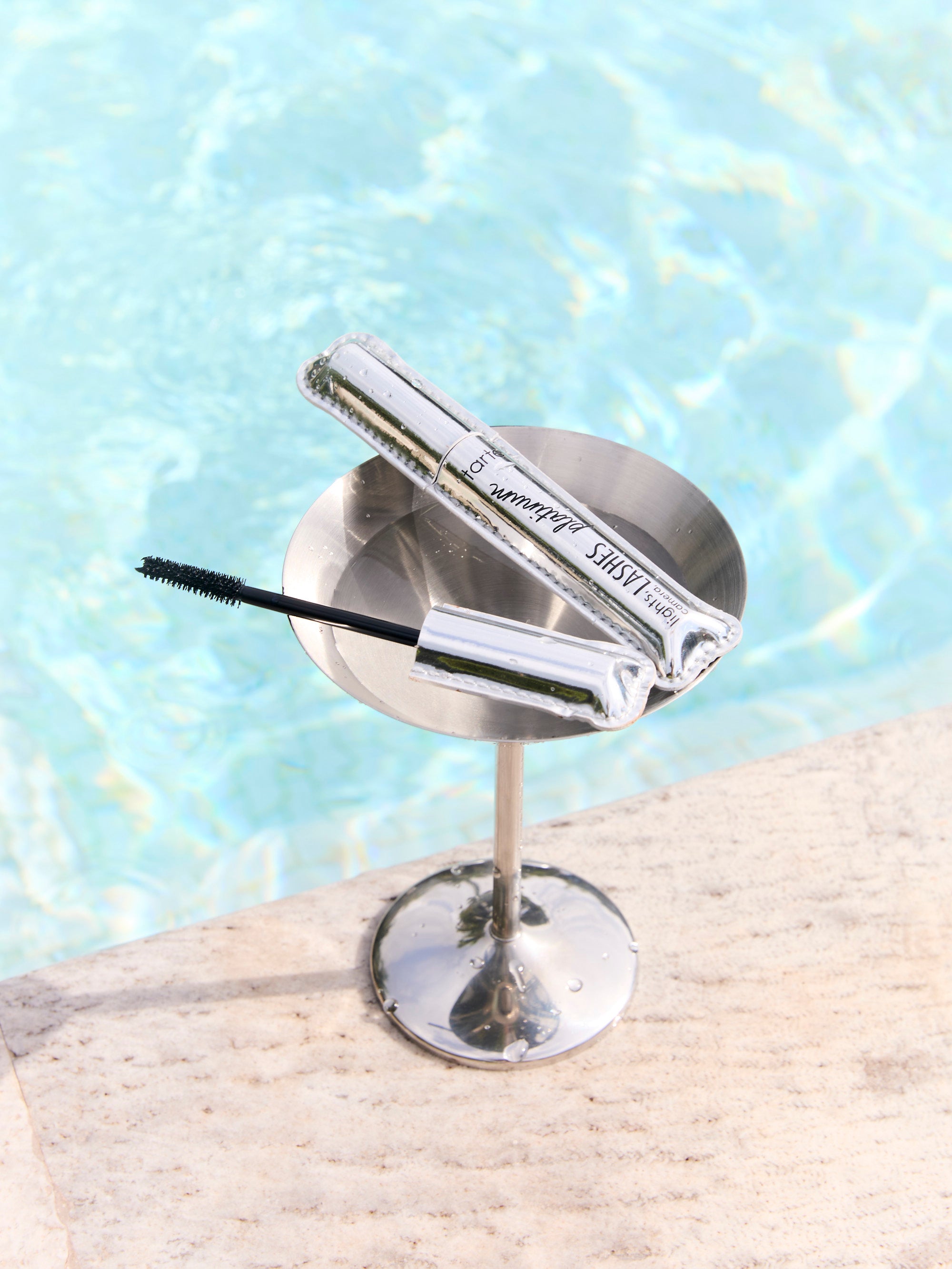 Mascara tube and wand rest on a metal stand beside a shimmering pool. Text on tube: tarte lights, camera, lashes 4-in-1 mascara.