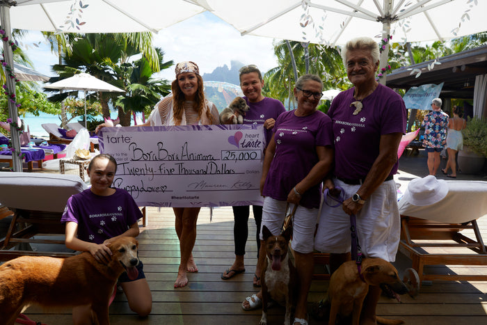 A group of people hold a large check to Bora Bora Animal Shelter for $25,000, on a deck with dogs around, under umbrellas, and by greenery and water. Text: tarte heart tarte.