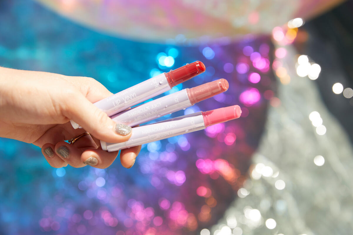 A hand holds three Tarte Cosmetics Maracuja Juicy Lip Vinyls in shades of red, pink, and peach against a colorful, sparkling background.