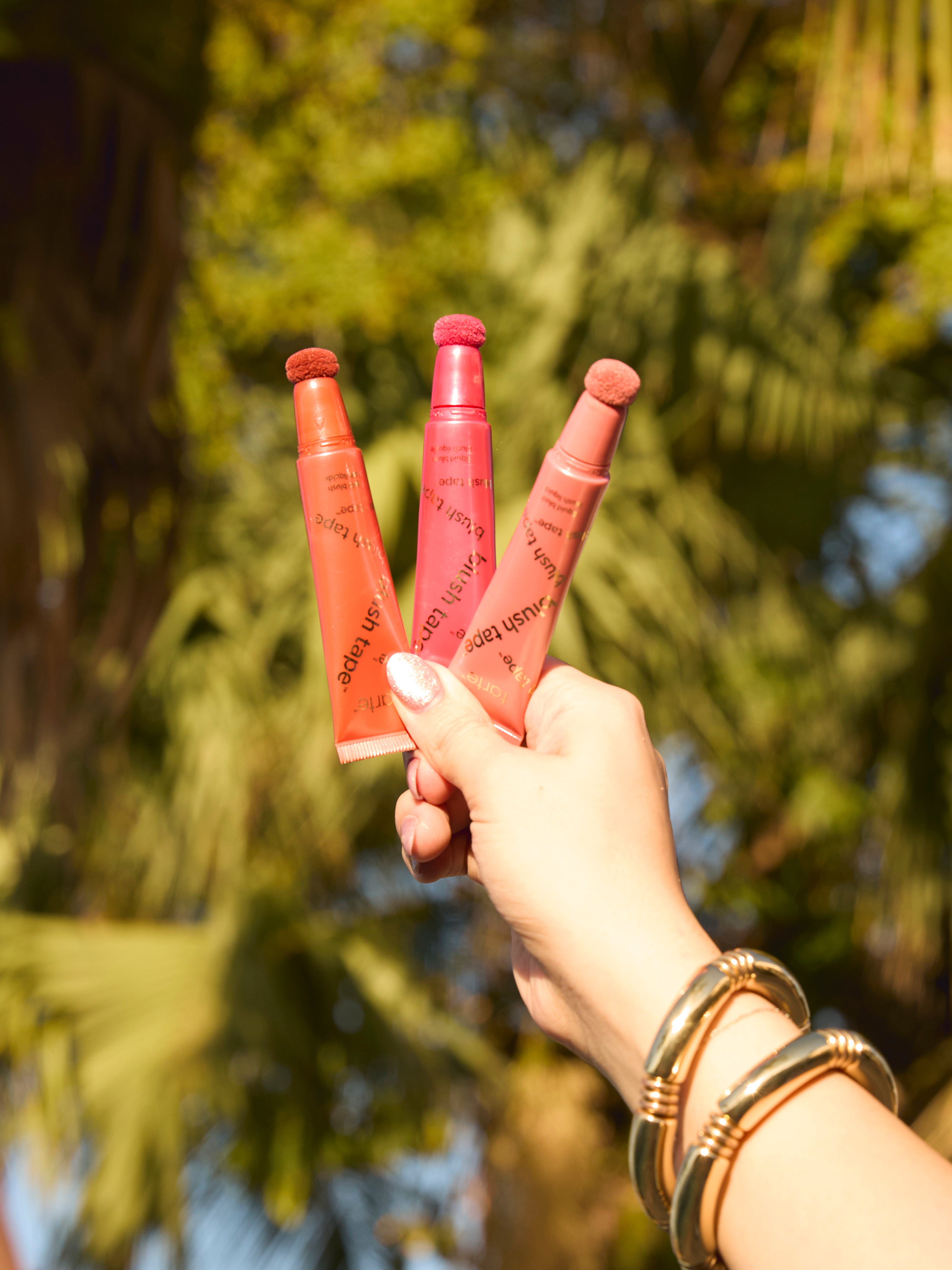 Three tubes of Tarte Cosmetics blush are held up by a hand wearing bangles, against a background of sunlit greenery.