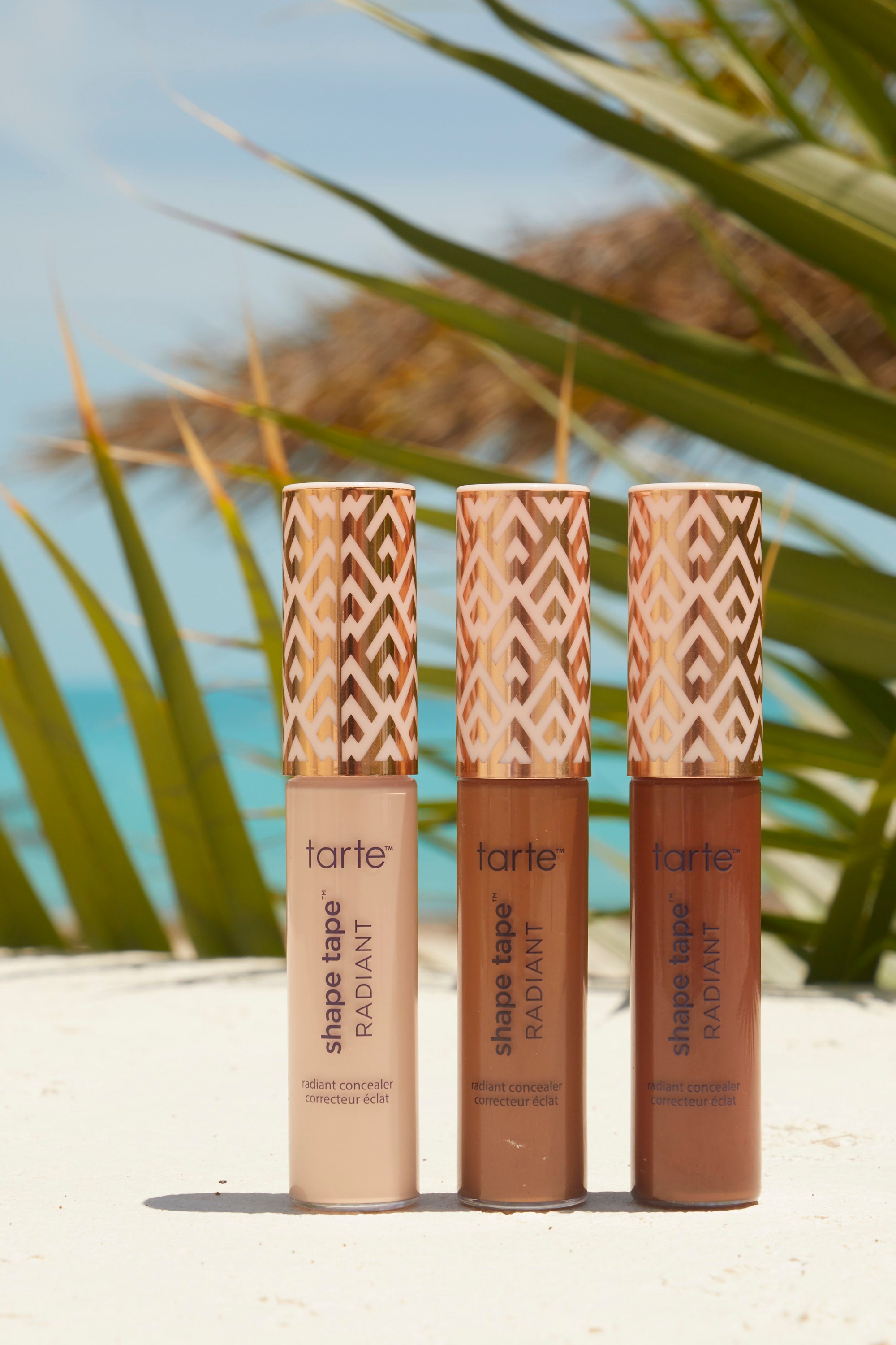 Three Tarte Cosmetics shape tape radiant concealers stand upright on a sandy surface. Palms and ocean in background. Text on bottles: shape tape™ RADIANT radiant concealer correcteur éclat.