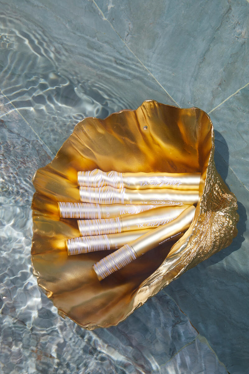 Golden shell holds multiple Tarte Cosmetics mascara tubes, text Tartelette XL, floating on clear water with rippling waves.