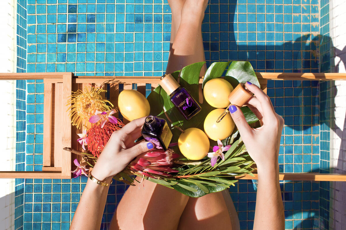 Hands hold Tarte Cosmetics maracuja oil dropper over fruits and flowers on a bamboo tray by a pool, with legs immersed in blue-tiled water.