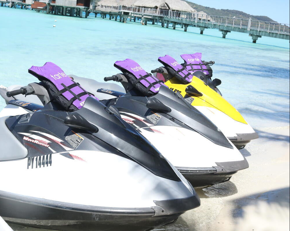 Jet skis with purple Tarte Cosmetics life jackets are lined up on a sandy beach near turquoise water. A pier with thatched-roof huts extends into the water in the background.