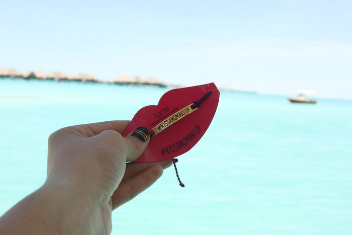 A hand holds a red, lip-shaped card with Tarte and KISSANDMAKEUP printed, featuring a small makeup item. The background shows turquoise water and distant, blurred huts on a sunny day.