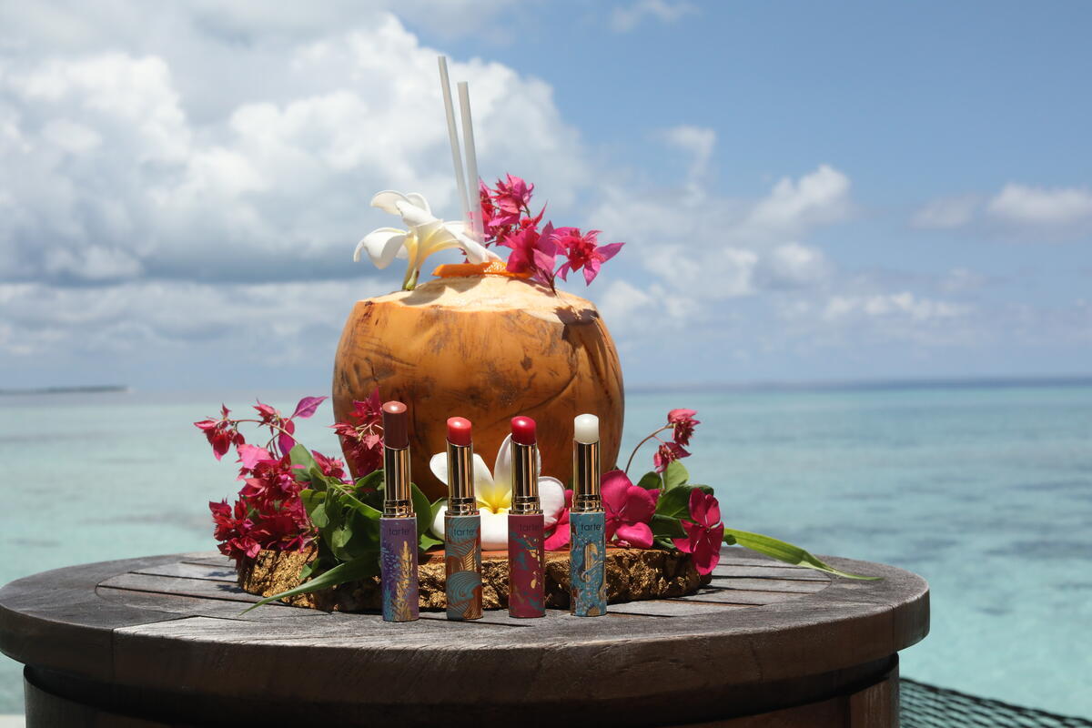 Lipsticks stand upright amidst bright flowers on a wooden table. A coconut with straws and more flowers sits behind, overlooking a tranquil ocean under a partly cloudy sky.