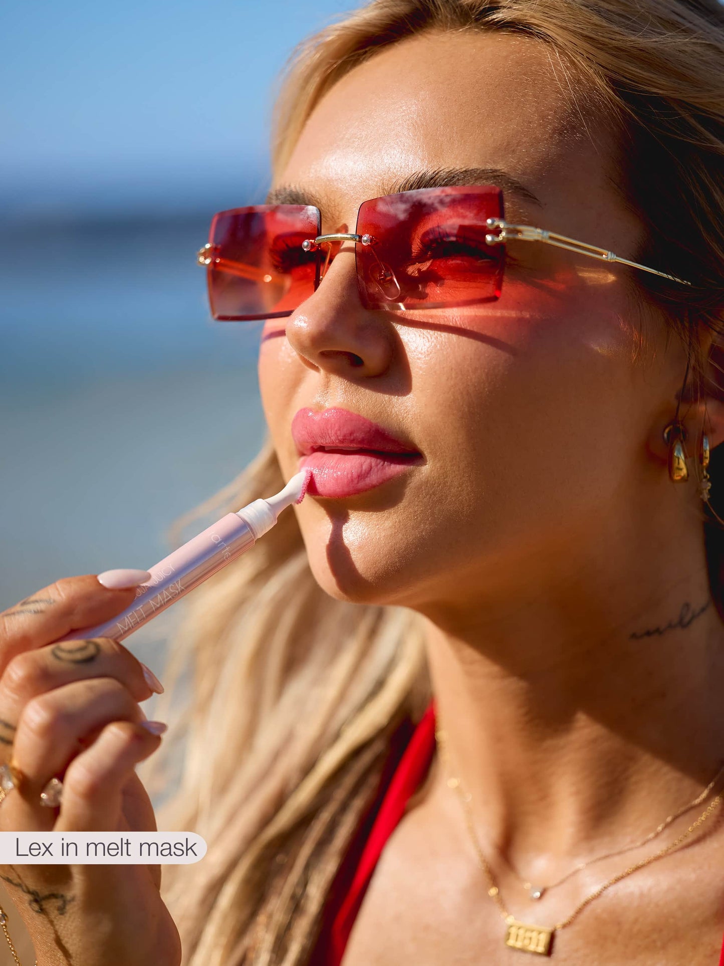 A person with long hair applies Tarte Cosmetics Melt Mask to their lips, wearing pink-tinted sunglasses and gold jewelry, set against a blurred outdoor background. Text: Lex in melt mask.
