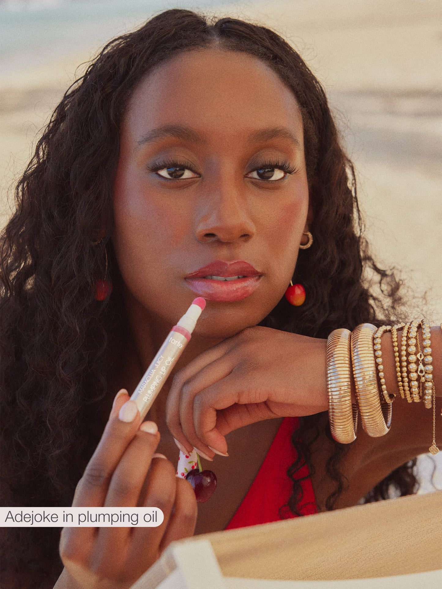 A person holds a tube of Tarte Cosmetics plumping lip oil, applying it to their lips. They're outside, possibly on a beach, wearing red attire and gold bangles. Text reads: Adejoke in plumping oil.