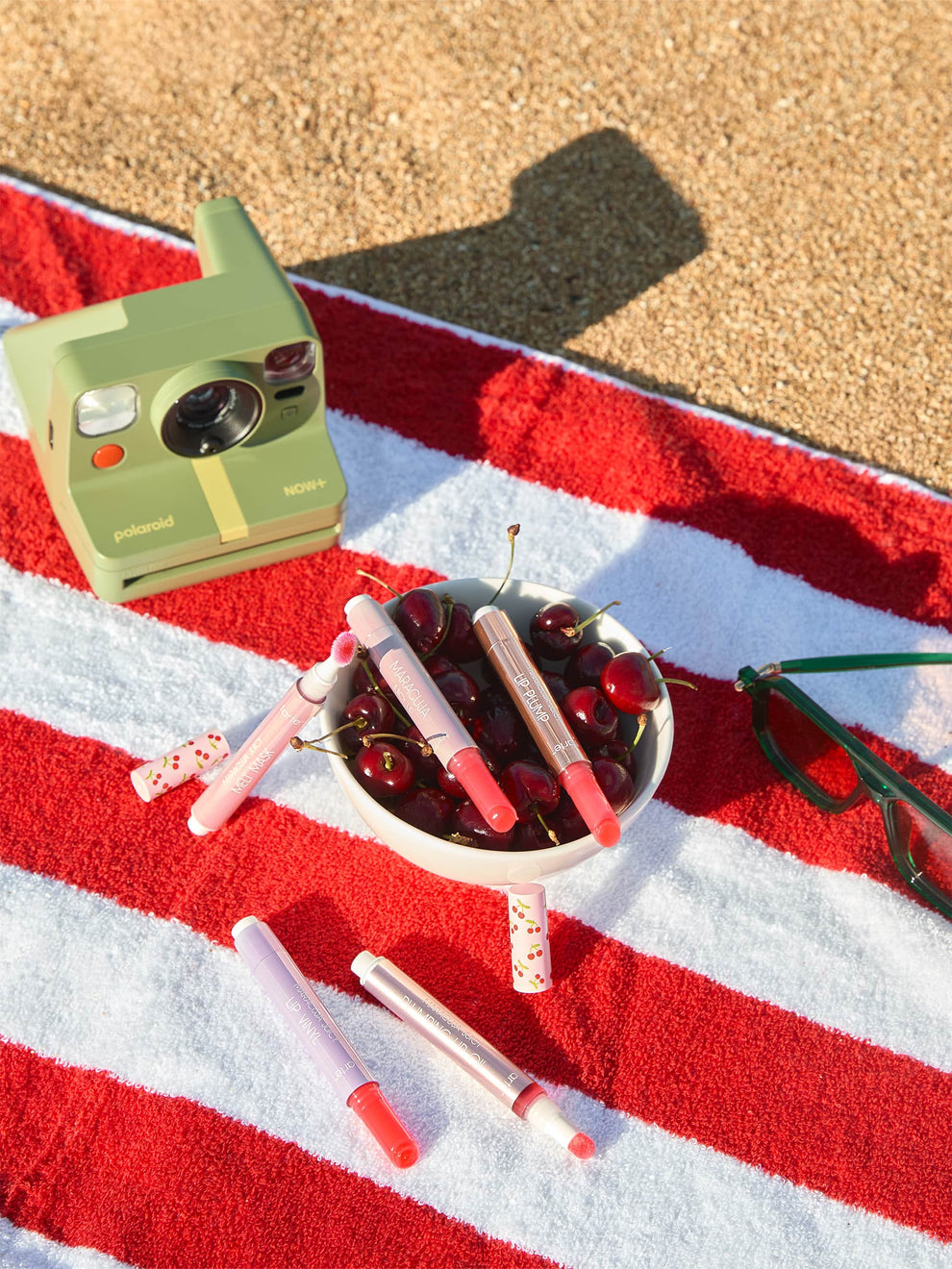 Tarte Cosmetics lip products are scattered across a red and white striped towel beside a bowl of cherries. Nearby, a green Polaroid camera and green sunglasses rest on the sandy surface.