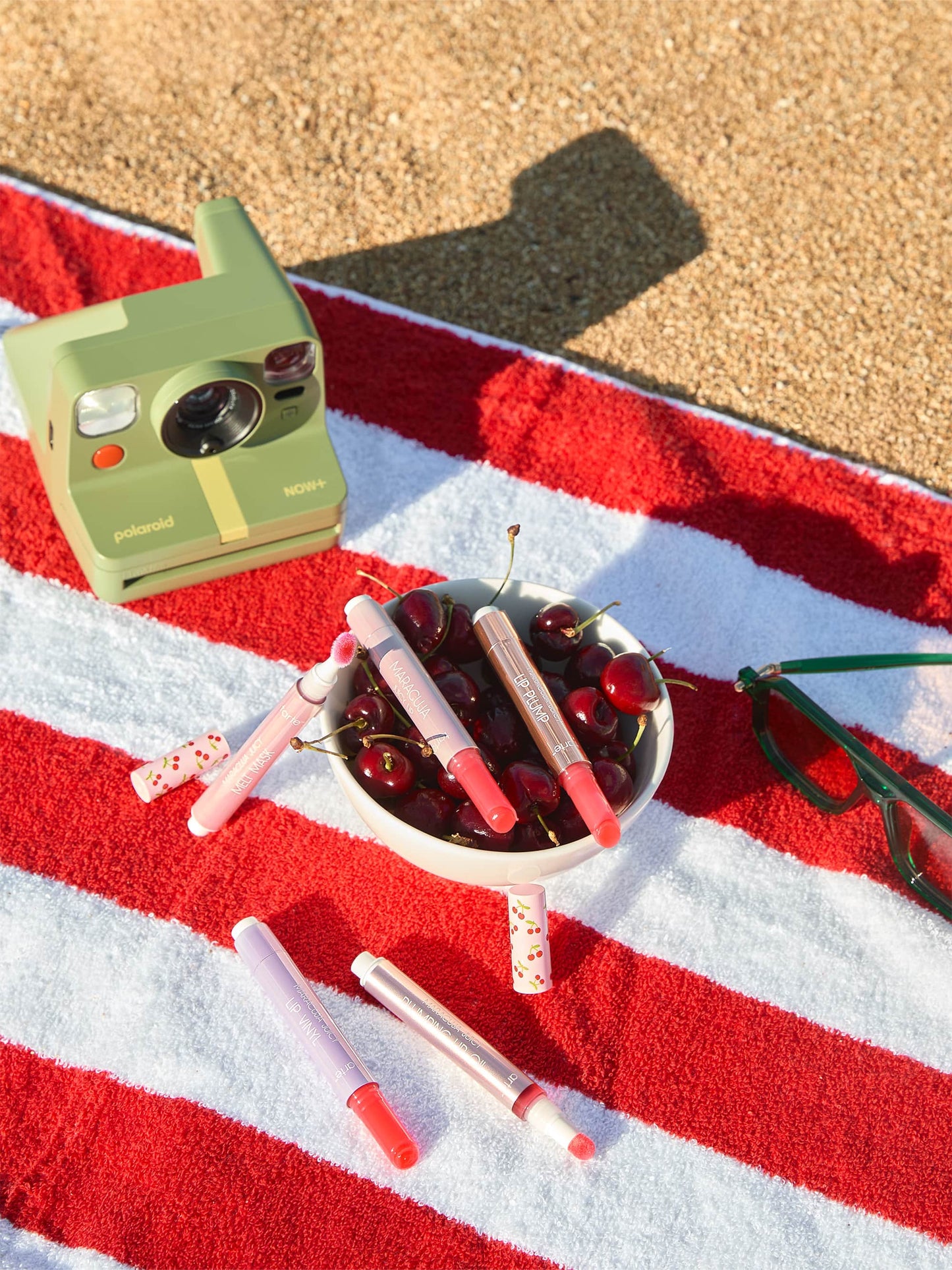 Tarte Cosmetics lip products are scattered across a red and white striped towel beside a bowl of cherries. Nearby, a green Polaroid camera and green sunglasses rest on the sandy surface.