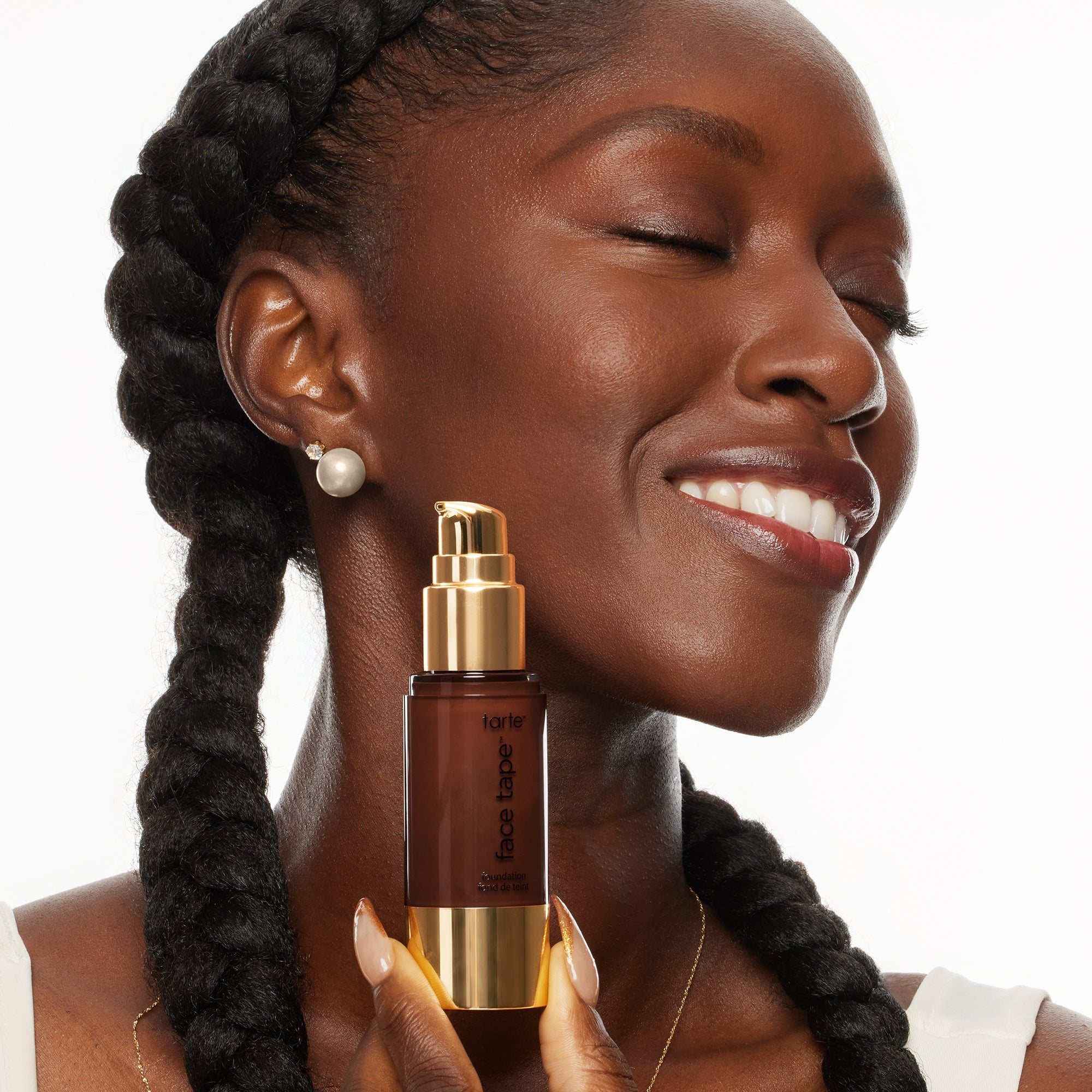 A person with braided hair smiles with eyes closed, holding a Tarte Cosmetics Face Tape foundation bottle with a gold pump in a bright, neutral background.