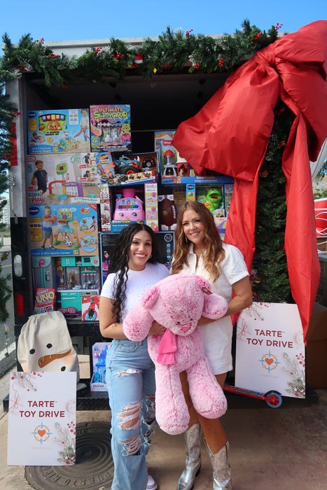 Two individuals holding a large pink teddy bear in front of a truck filled with toys, adorned with a large red bow and green garlands. Signs read Tarte Toy Drive.