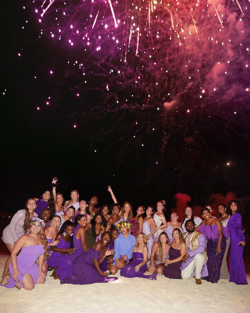 A group of people, mostly in purple attire, pose joyfully on a beach at night, with colorful fireworks illuminating the dark sky above them.