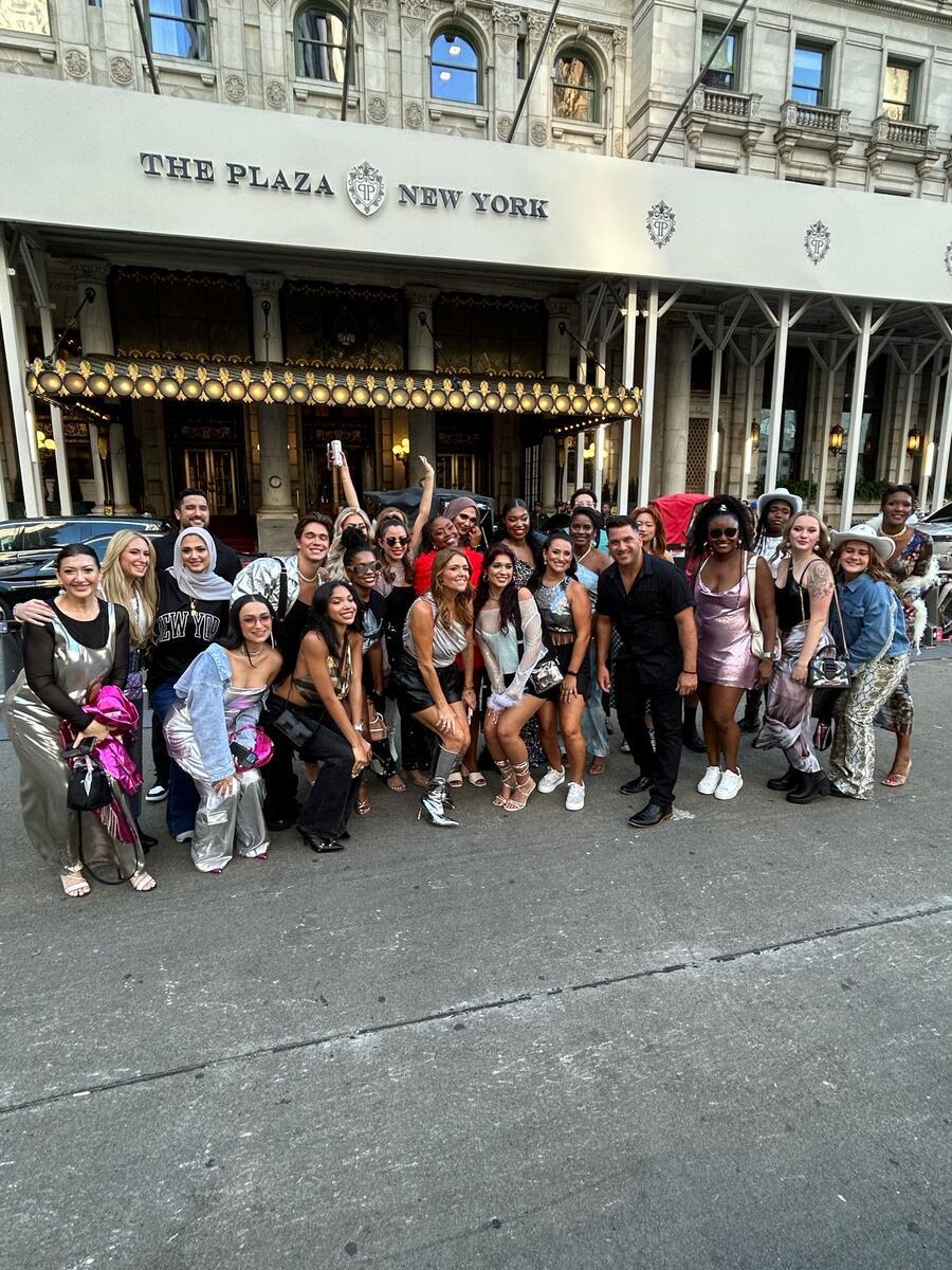 A group of people gather, smiling and posing on a street, in front of the entrance to The Plaza New York. The building's ornate facade and signage are visible above them.