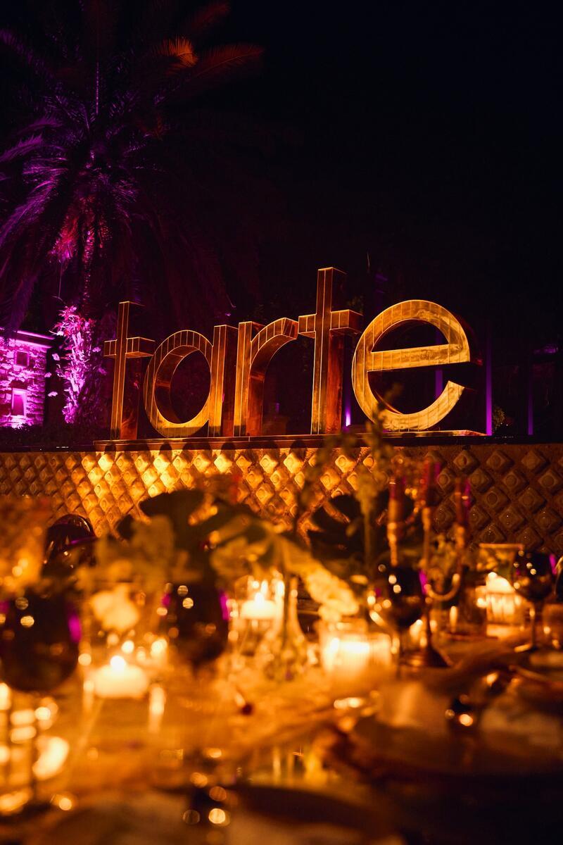 Illuminated letters spell tarte beside a decorative table with candles and flowers. A lit palm tree stands in the background, creating a warm, festive atmosphere.