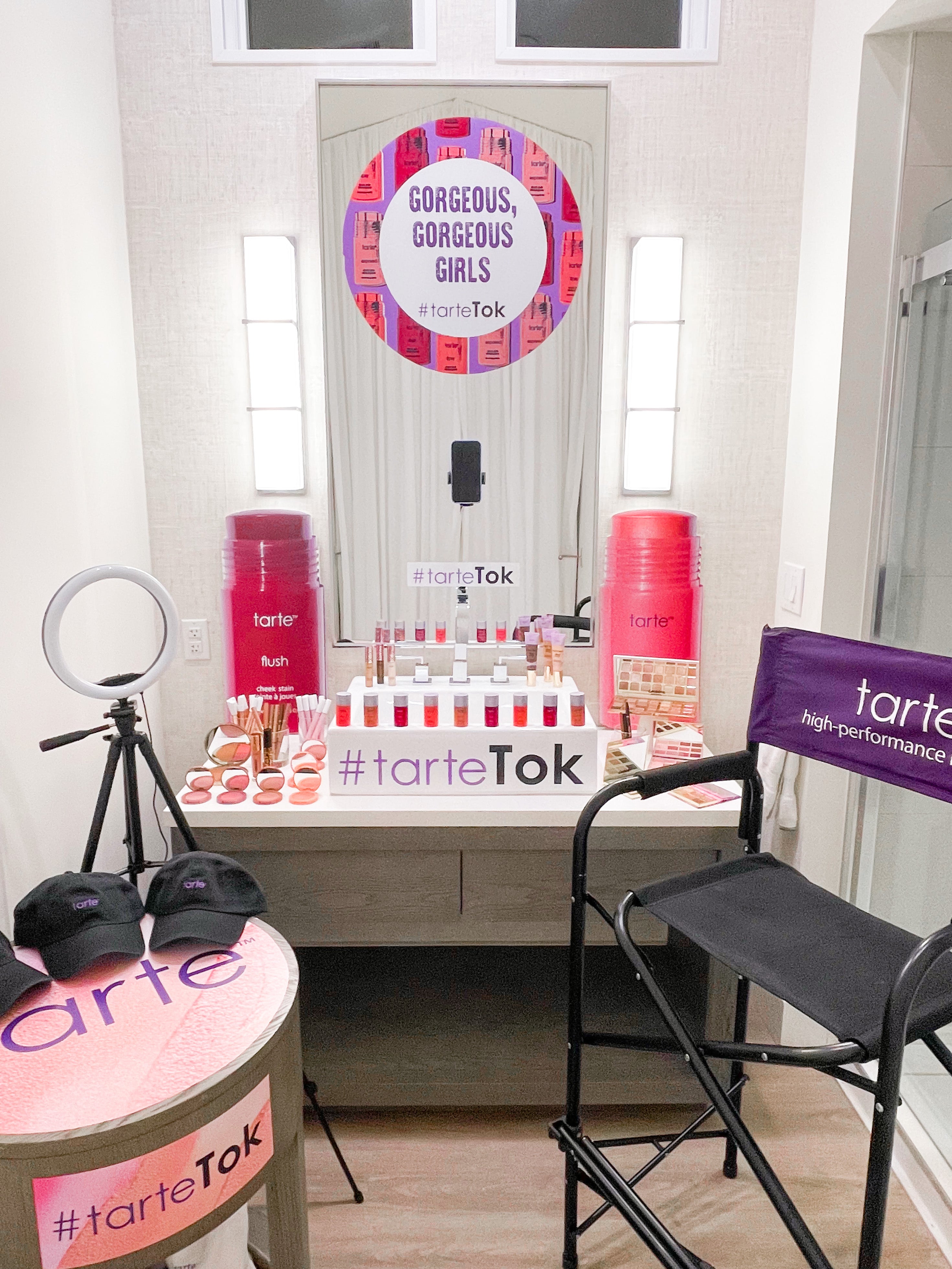 A makeup display, featuring Tarte Cosmetics products, sits on a vanity under a sign reading GORGEOUS, GORGEOUS GIRLS tarteTok. Surrounding context includes a ring light, branded chair, and hats.