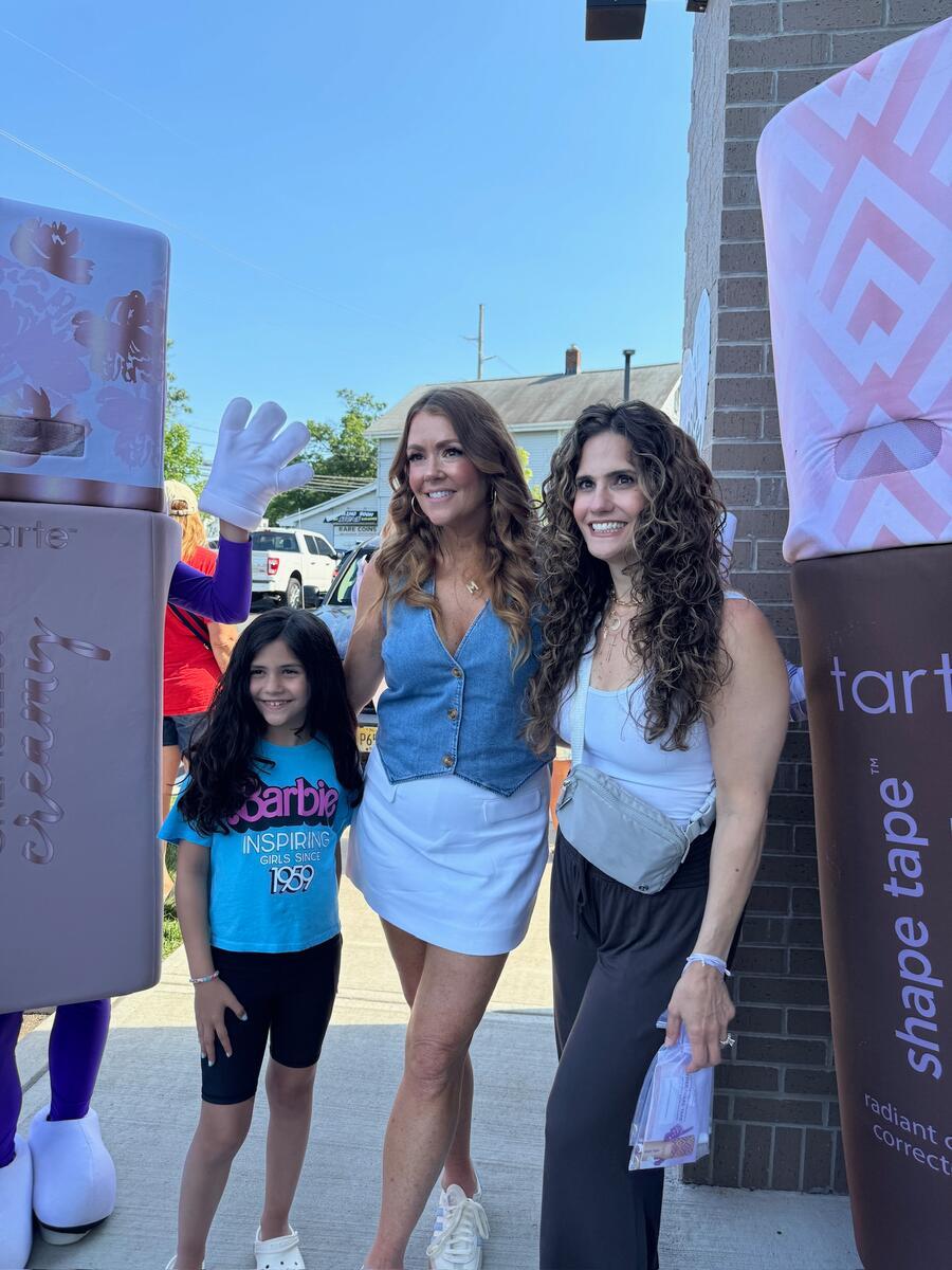 Three people stand smiling outside next to Tarte Cosmetics' large product displays. A girl in a Barbie Inspiring Girls Since 1959 shirt is on the left. A brick building and blue sky are in the background.