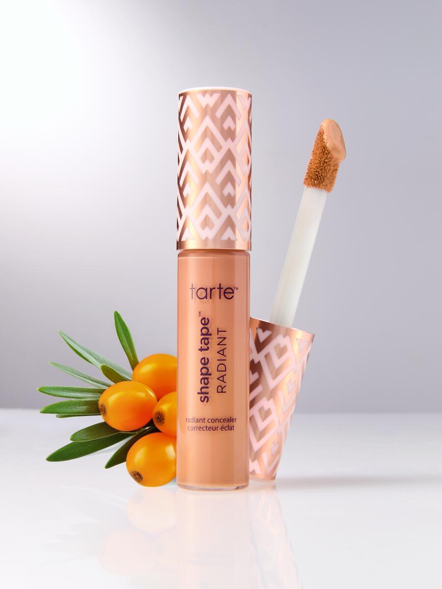 A Tarte Cosmetics Shape Tape Radiant concealer with an applicator wand, displayed upright beside small orange berries and green leaves on a reflective surface.