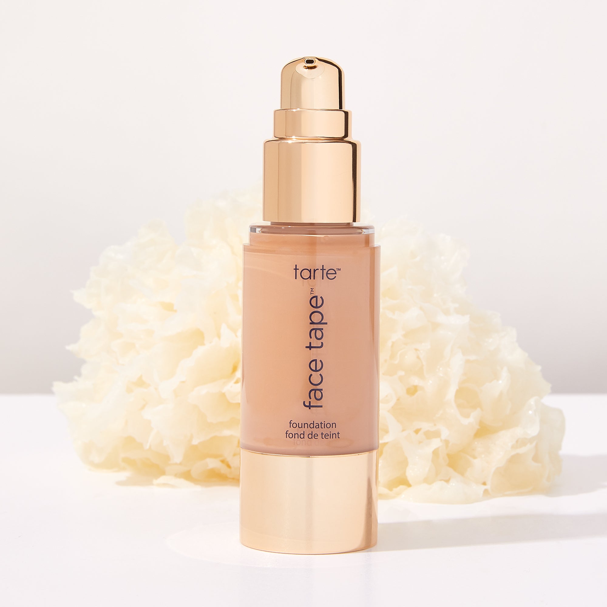A bottle of Tarte Cosmetics Face Tape foundation stands upright, featuring a golden pump, against a backdrop of white, textured floral decorations. Text reads: tarte™ face tape™ foundation fond de teint.