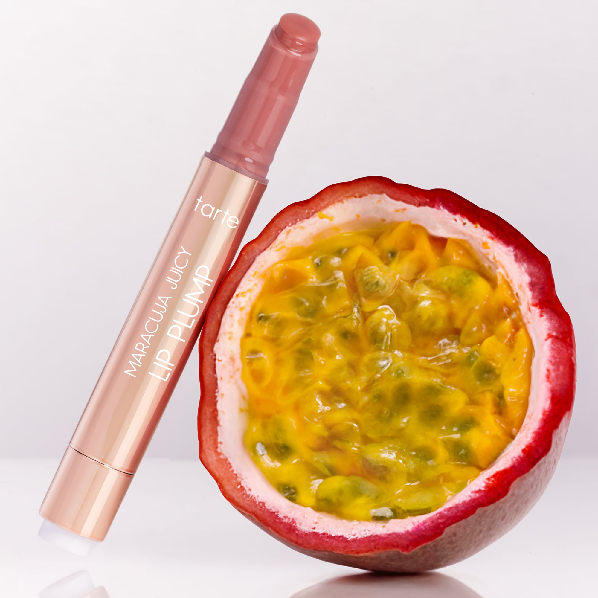 A Tarte Cosmetics Maracuja Juicy Lip Plump stands upright beside a halved passion fruit, displaying vibrant yellow seeds. The background is a clean, white surface.