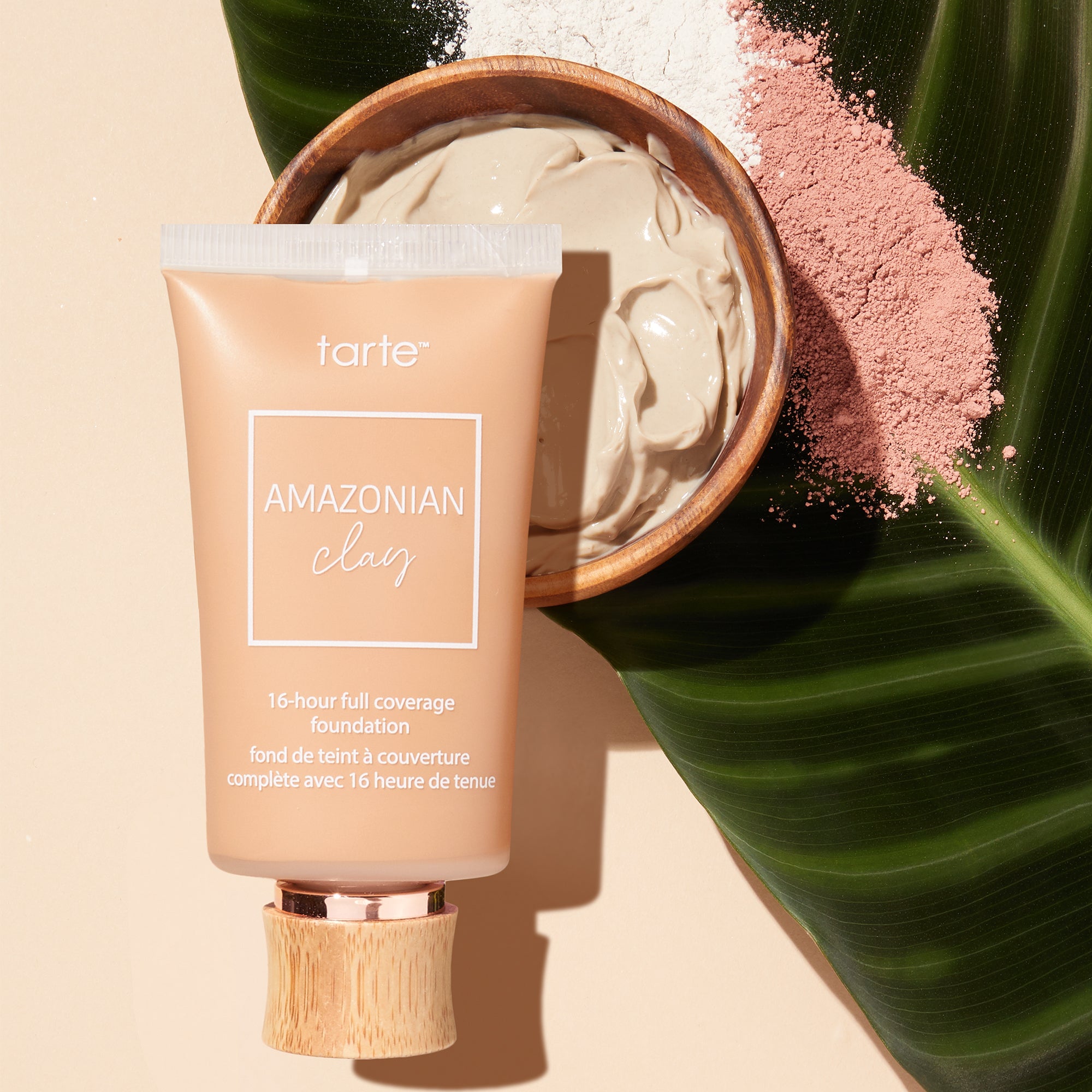 A tube labeled tarte AMAZONIAN clay, 16-hour full coverage foundation rests on a tropical leaf. Nearby, a wooden bowl holds clay, surrounded by pink and white powders.