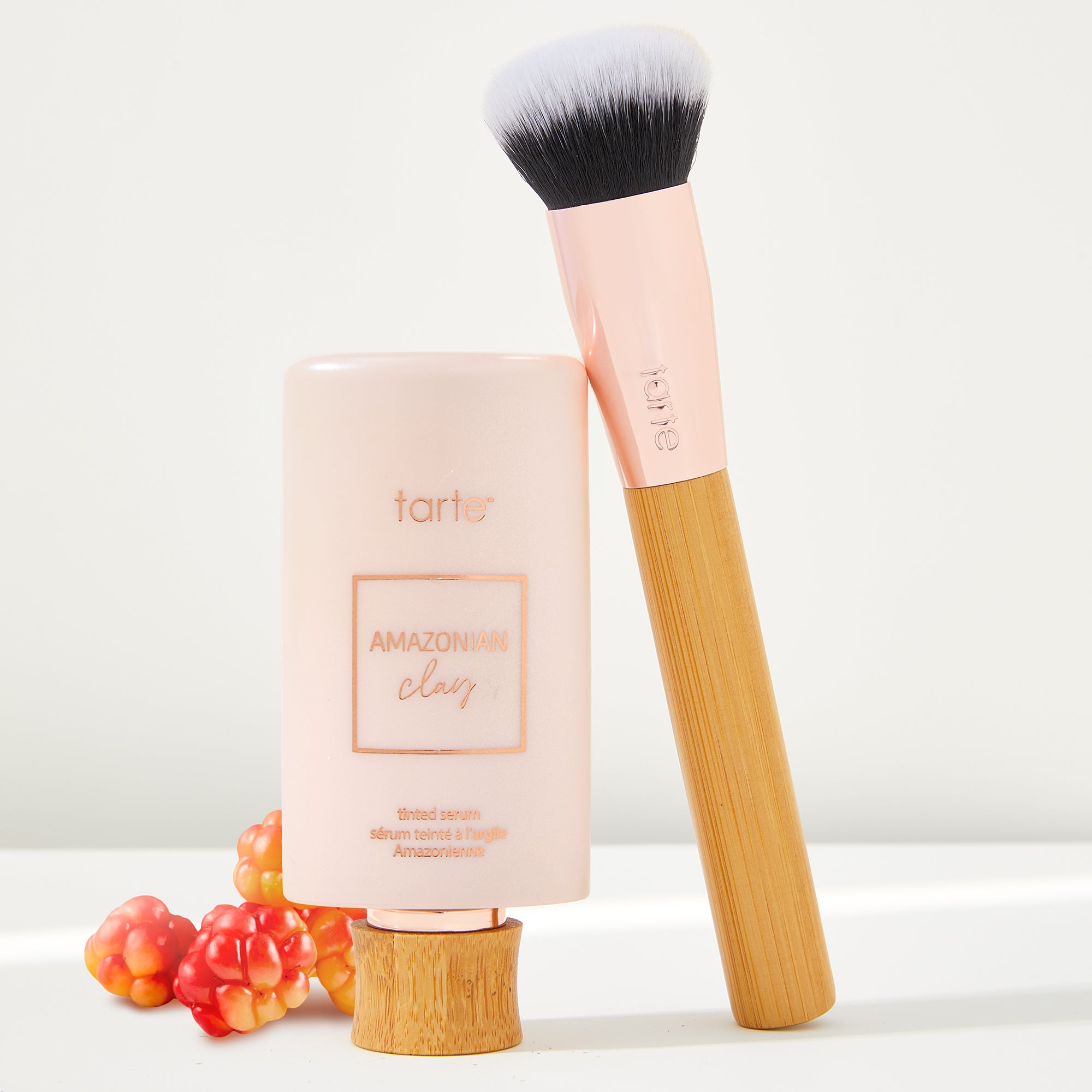 A foundation bottle labeled tarte Amazonian clay tinted serum stands upright, accompanied by a makeup brush with a wooden handle. Small orange berries lie nearby against a plain background.
