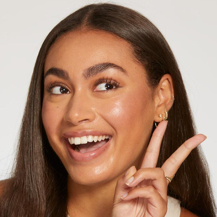 A smiling person holds up a peace sign, showcasing glowing skin and wearing gold hoop earrings, with smooth, straight hair against a plain white background.