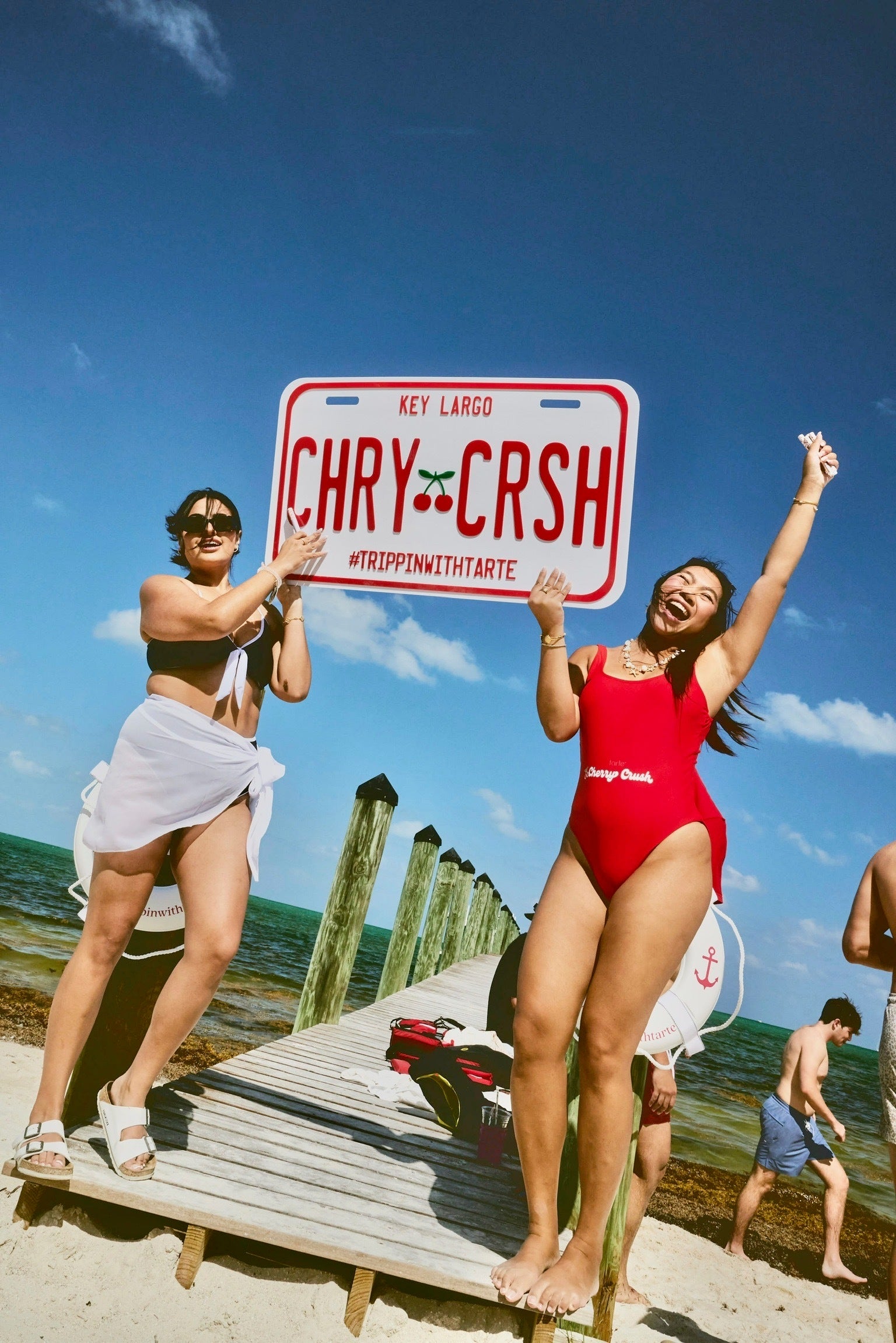 Two individuals hold a large sign reading Key Largo CHRY-CRSH TRIPPINWITHTARTE, standing on a sandy beach with a wooden dock extending into calm, turquoise water.