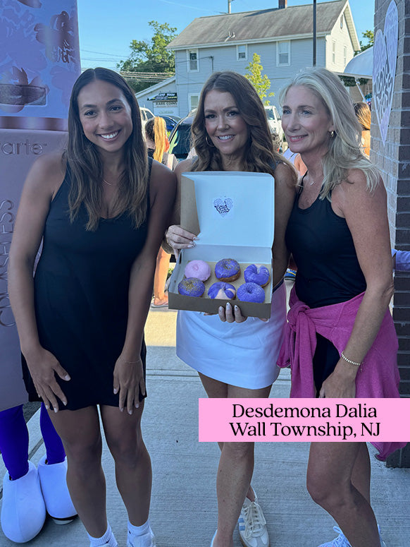 Three women stand outdoors holding a box of colorful donuts. They are smiling, dressed casually in black outfits, with a building and people in the background. Text: Desdemona Dalia, Wall Township, NJ.