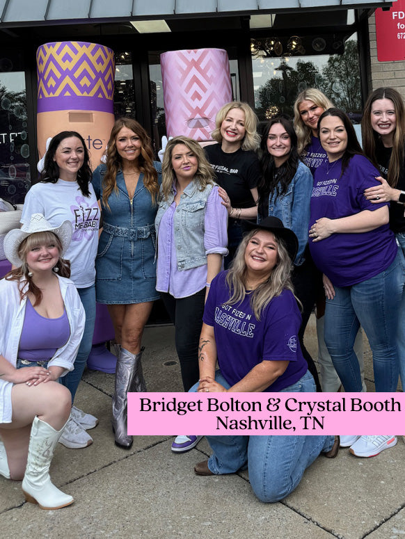 A group of eleven women, some wearing purple and casual outfits, pose outside a storefront featuring large Tarte Cosmetics displays. Text: Bridget Bolton & Crystal Booth, Nashville, TN.