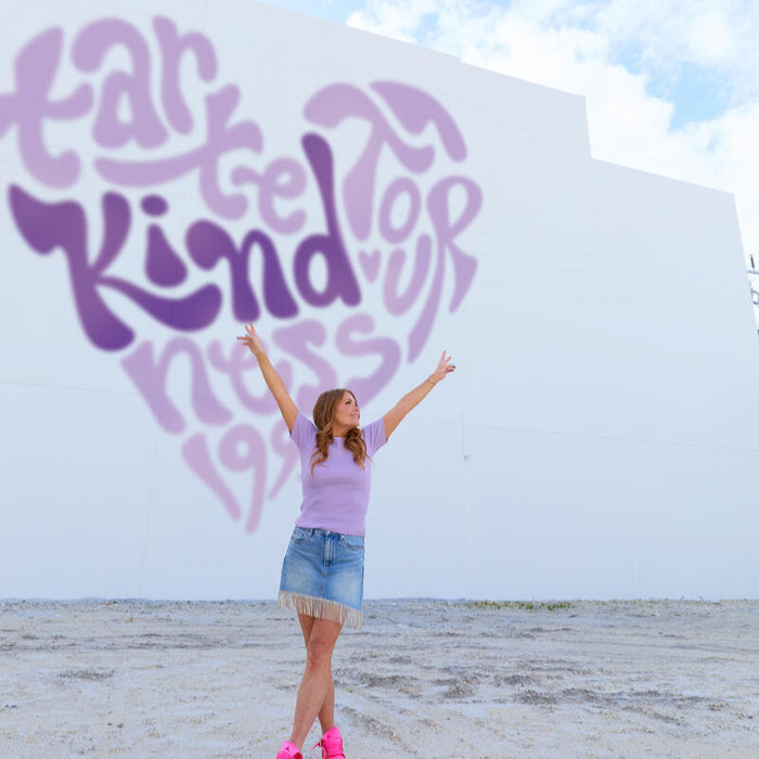 A person stands with arms raised on a sandy ground before a white wall featuring large purple text: tarte top kindness 199. The sky above is partly cloudy.