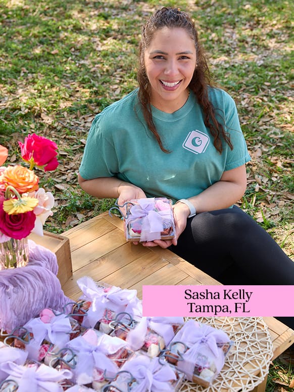 A person wearing a green shirt smiles while holding a decorated box outdoors. A wooden table displays more boxes and vibrant flowers. Text reads: Sasha Kelly, Tampa, FL.
