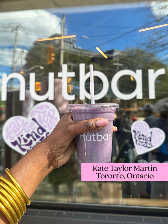 A hand holds a purple smoothie in front of a glass window with nutbar written on it. Background includes street reflections. Text: Kate Taylor Martin, Toronto, Ontario.