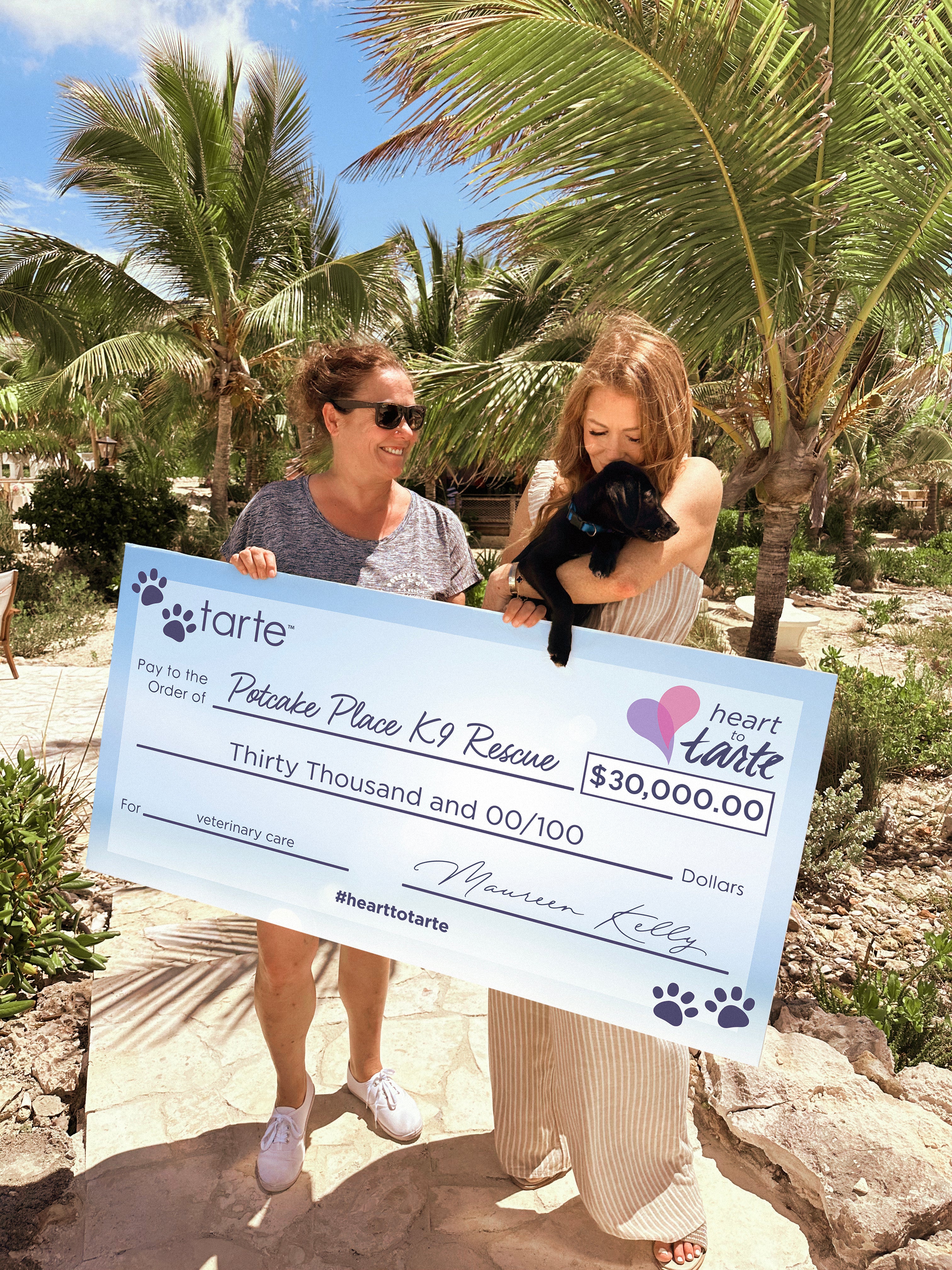 Two people hold a giant check for $30,000 made out to Potcake Place K9 Rescue from Tarte Cosmetics, standing on a sunlit path with palm trees. One holds a black puppy.