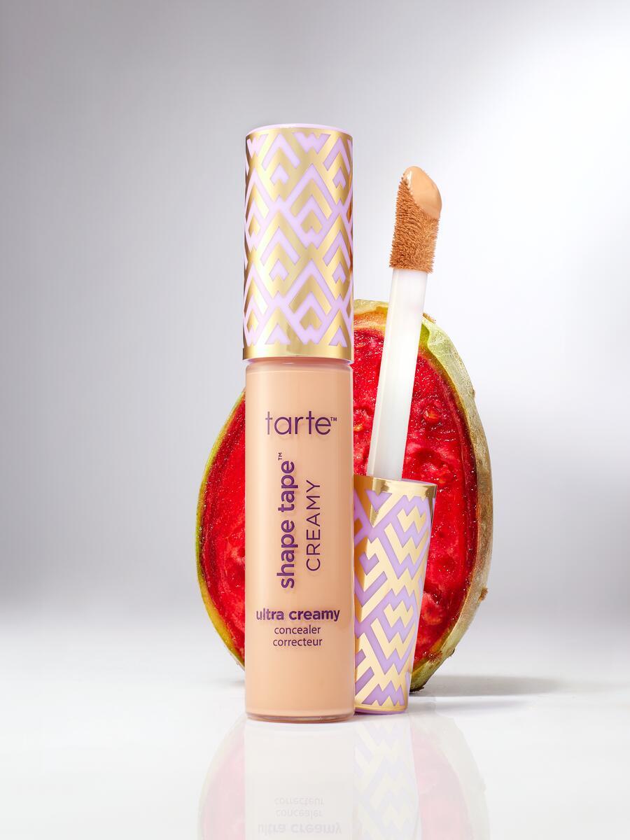 A Tarte Cosmetics Shape Tape Creamy concealer stands next to a half watermelon. The concealer's applicator is visible, resting against its decorative cap. Text on the bottle reads: ultra creamy concealer correcteur.