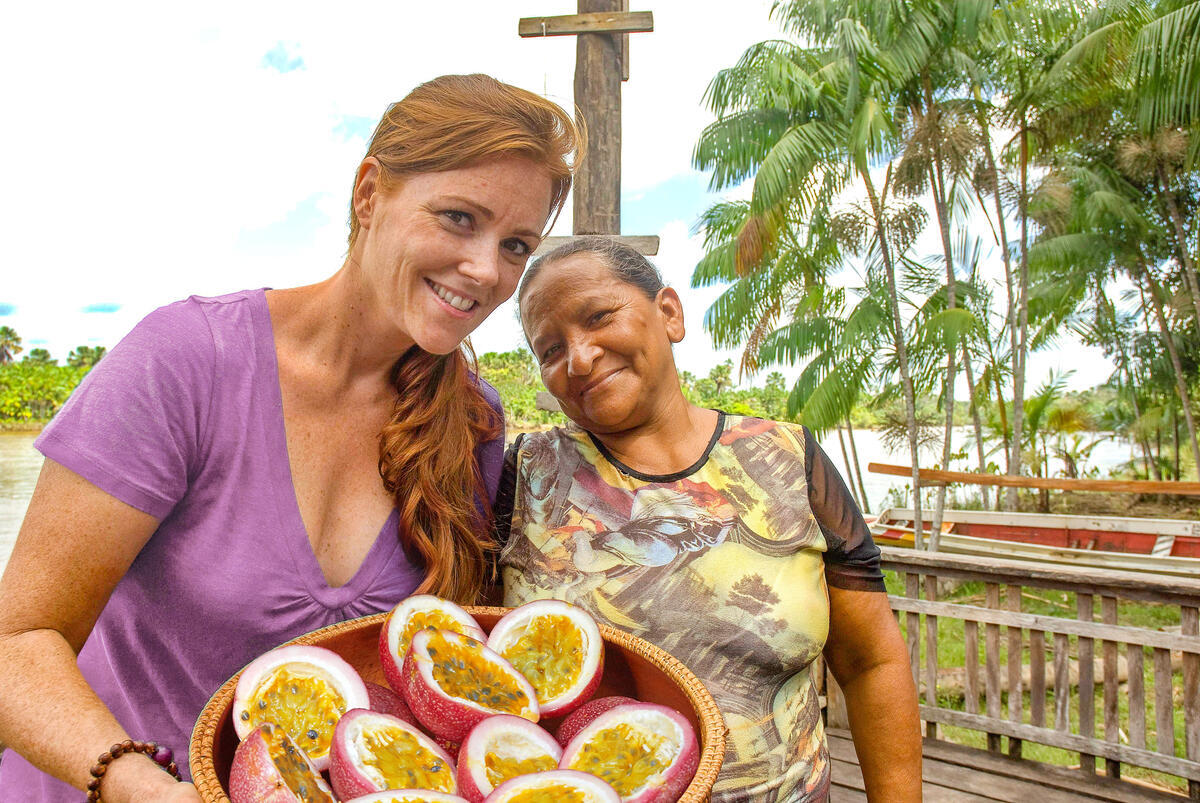Two women smiling outdoors, one holding a bowl of halved passion fruits. They're on a wooden deck, surrounded by lush greenery and palm trees under a partly cloudy sky.