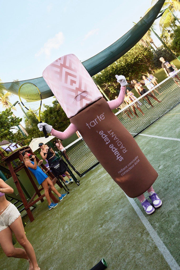A person in a Tarte Cosmetics concealer tube costume holds a tennis racket on a tennis court, surrounded by spectators and greenery. The costume reads: tarte® shape tape™ RADIANT radiant concealer correcteur éclat.
