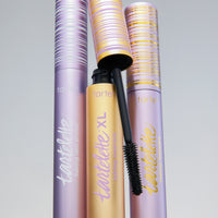 Three tubes of Tarte Cosmetics products stand upright. The central tube is open, showcasing a mascara brush. Purple and gold packaging features tartelette branding with text: tubing lash primer and tubing mascara XL.