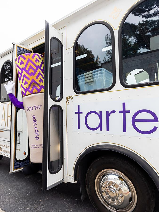 A large, costume character resembling Tarte Cosmetics' Shape Tape exits a branded bus on a road, with surrounding trees and storage boxes visible inside.