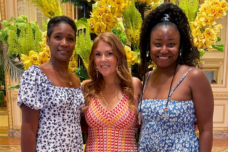 Three women stand smiling, wearing summer dresses in a lavish indoor setting with yellow flowers and greenery surrounding them. They appear relaxed and happy together in the ornate environment.