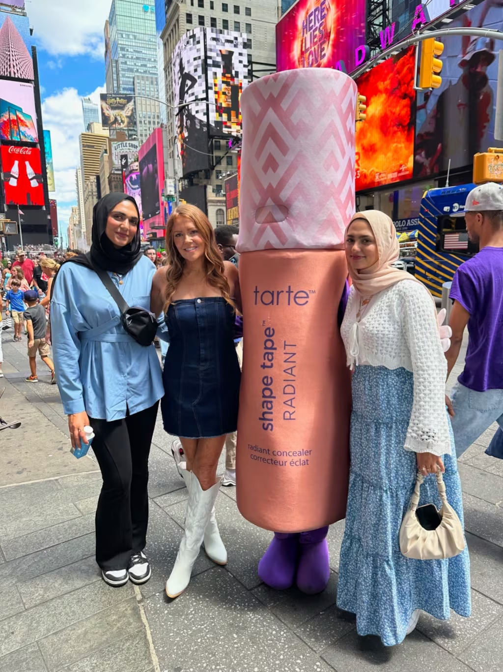A giant Tarte Cosmetics concealer tube costume is surrounded by three women in Times Square. Skyscrapers and colorful billboards tower above, creating a bustling urban backdrop.
