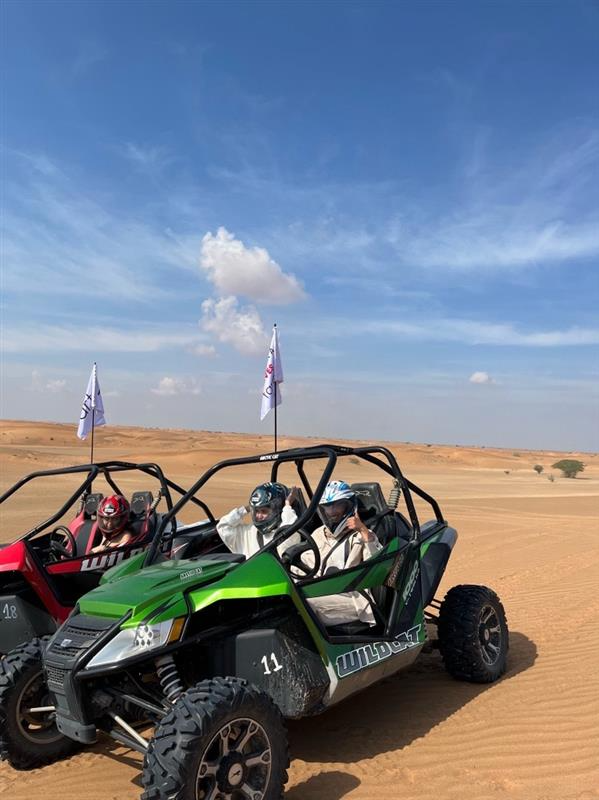 Two dune buggies, one green and one red, carry helmeted passengers across a sandy desert landscape under a clear blue sky with scattered clouds. White flags are mounted on the buggies.