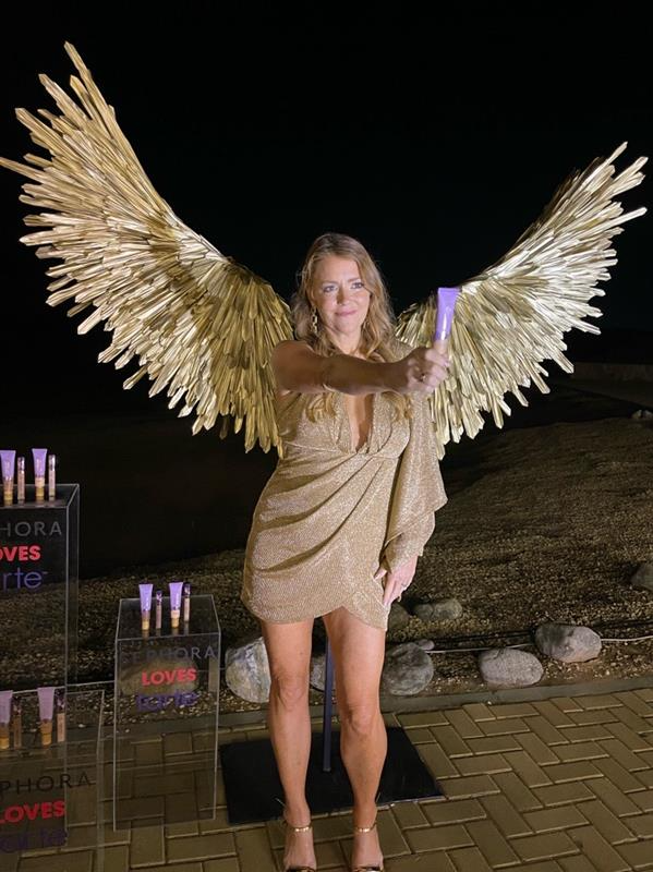 A woman in a gold dress, with large golden wings, holds a purple tube in one hand. She stands outdoors on a brick path at night. Text: SEPHORA LOVES tarte.