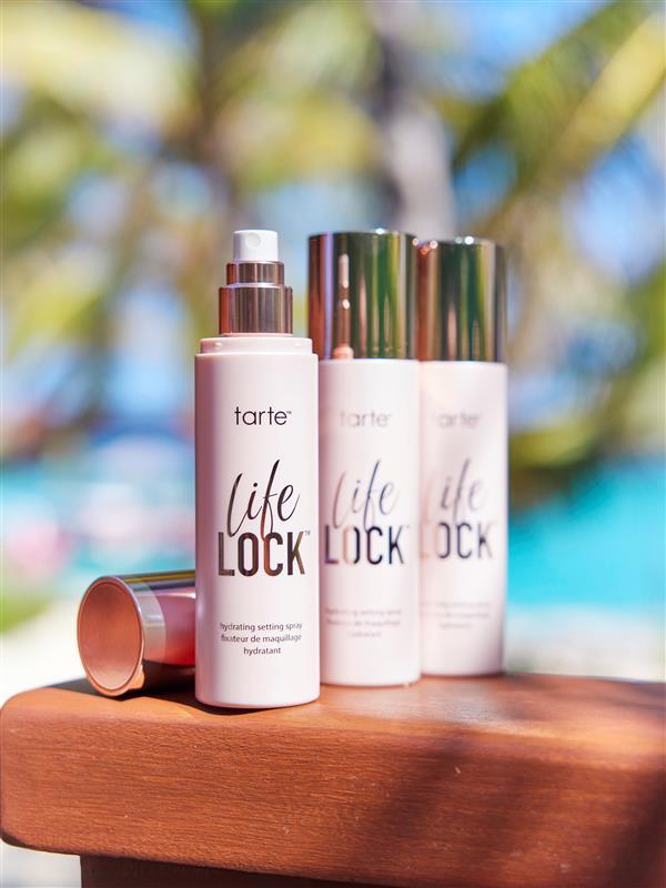 Bottles of Tarte Cosmetics Life Lock hydrating setting spray are standing on a wooden surface, with palm trees and a vibrant blue sky in the blurred background.