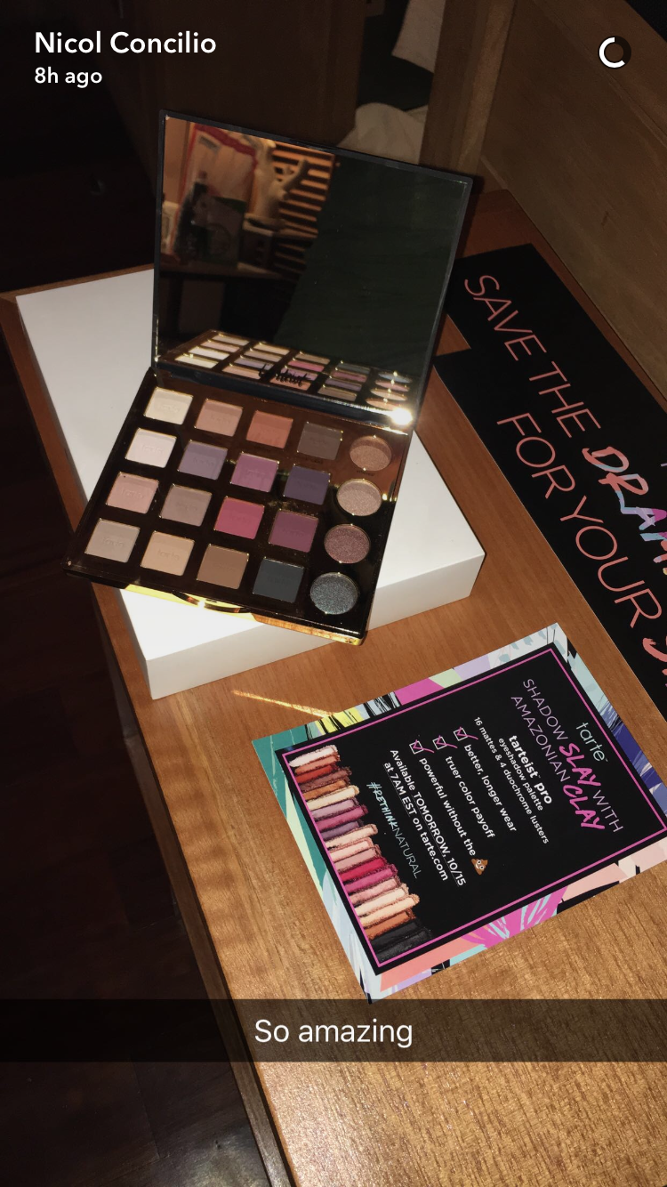 A Tarte Cosmetics eyeshadow palette on a table reflects light with open display. Nearby, promotional cards detail product features, stating Tarte Slay with Amazonian Clay. Text overlay: So amazing.