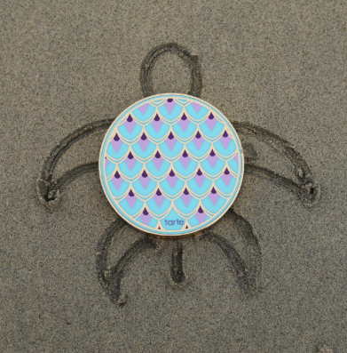 Compact with blue and pink scale pattern rests on sand, centered in a turtle drawing. The environment is a beach setting. Text: tarte.