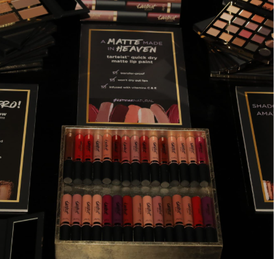 A display shows rows of Tarte Cosmetics quick dry matte lip paint tubes in various shades, surrounded by makeup products. Signs highlight features: waterproof, won't dry out lips, infused with vitamins C & E.